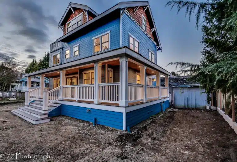 A large blue house with white trim and a wraparound porch, nestled in West Seattle, is surrounded by bare dirt and trees, photographed at dusk.