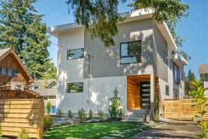 Modern two-story house with large windows, light gray siding, white panel accents, and a wooden entryway. The front yard features green grass, small shrubs, and a wooden fence.