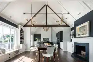 Bright, modern dining room with vaulted ceiling, exposed beams, contemporary light fixtures, large windows, a fireplace with artwork above, and a wooden table with white chairs.