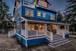 A modern blue three-story house with white trim, lit windows, and a wraparound porch sits nestled in West Seattle. Wide steps lead to the main entrance. Surrounded by trees, it’s dusk outside.