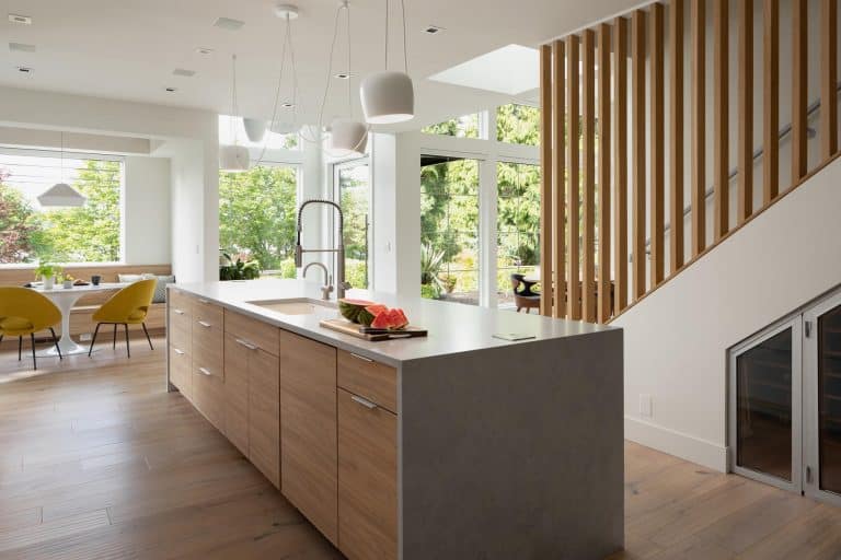Modern kitchen with wooden island, white countertops, pendant lights, and a view of a dining area with yellow chairs by large windows overlooking green trees. Stairs with vertical wooden slats are on the right.
