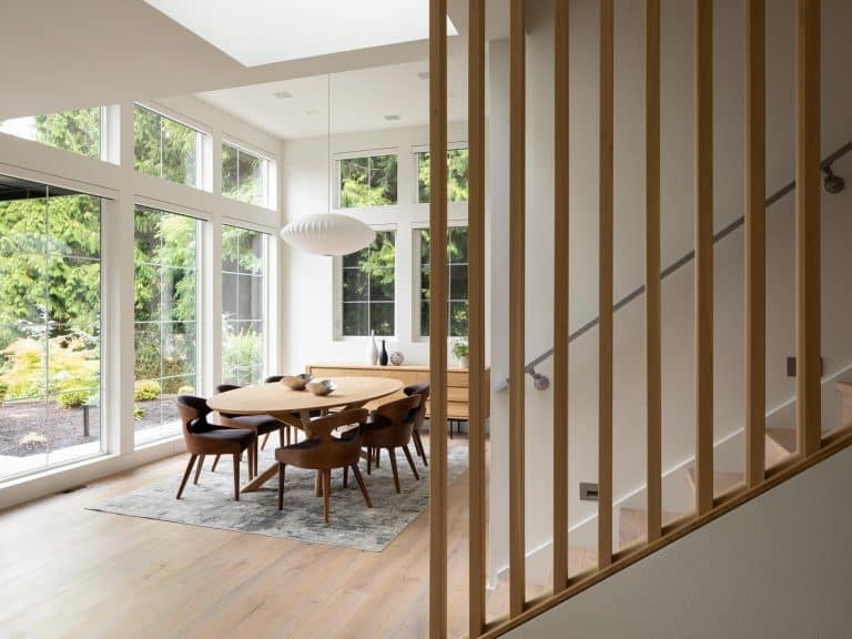 Modern dining room with large windows, wooden table and dark chairs, light wood floors, a pendant light, and a staircase with vertical wooden slats; lush greenery is visible outside.
