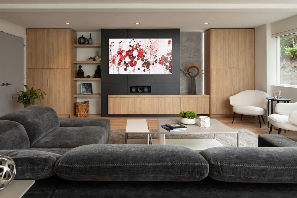 Modern living room with gray sofas, a large TV displaying floral art, wooden cabinets, white armchairs, a marble coffee table with books and a plant, and natural light coming from a window on the right.