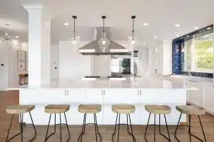 Modern kitchen with a large white island, five wooden bar stools with black metal legs, stainless steel appliances, glass pendant lights, and a big window with blue tile backsplash overlooking greenery.