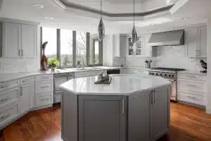 Modern kitchen with gray cabinets, white marble countertops, stainless steel appliances, a central island, pendant lights, and large windows letting in natural light. Decor includes plants and sculptures.