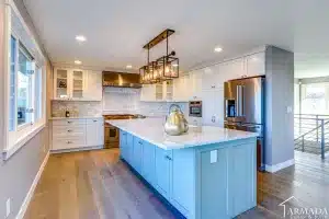 Bright modern kitchen with white cabinets, stainless steel appliances, a blue island with a marble countertop, gold kettle, wooden floors, and a geometric light fixture above the island.