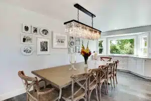 Bright dining room with a wooden table, six chairs, a modern chandelier, flower centerpiece, large windows, and framed photos on the wall.