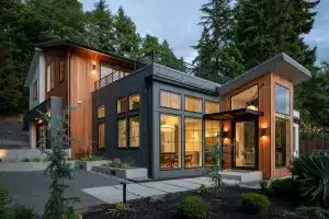 Modern two-story house with large windows, wood and dark panel siding, outdoor lights, and surrounded by trees and neatly landscaped plants at dusk—creating a setting as inviting and organized as a well-designed Services Menu.