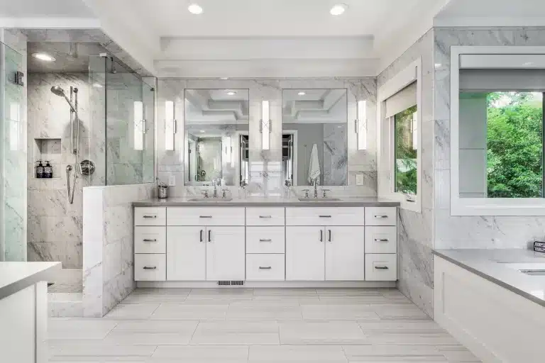 Modern bathroom with white cabinetry, double sinks, large mirrors, marble walls, glass shower, tiled floor, and a window with green trees visible outside. Bright lighting and a clean, sleek design.