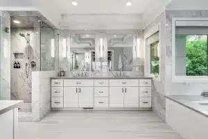Modern bathroom with white cabinets, double sinks, a large mirror, wall sconces, glass shower, marble walls, light tile floors, and a window showing green trees outside.