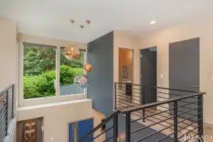Modern interior upper hallway with black metal railing, three hanging pendant lights, large window with a green outdoor view, and doors leading to other rooms. Light walls and natural lighting create an airy atmosphere.