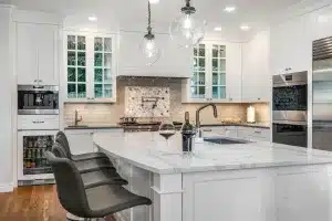 Modern kitchen with white cabinets, marble island, pendant lights, stainless steel appliances, three dark chairs, and two glasses of wine with a wine bottle on the counter. Light wood floors and a tiled backsplash are visible.