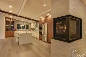Modern kitchen with wood floors, white cabinets, stainless steel appliances, large island with stools, and a corner fireplace. Recessed lighting and wood ceiling beams add warmth. ARMADA DESIGN & BUILD logo in the bottom right.
