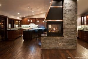 Modern kitchen with dark wood cabinets, island seating, and a double-sided stone fireplace. Hardwood floors and pendant lights create a warm, inviting atmosphere. Photo credit: Jesse L. Young Photography.