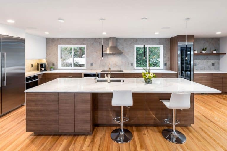Modern kitchen with wooden cabinets, stainless steel appliances, white countertops, two bar stools, pendant lights, and large island with sink. Hardwood floors and large windows provide natural light.