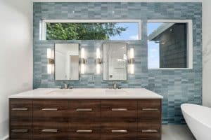 Modern bathroom with a double sink vanity, wood drawers, two mirrors, vertical lights, blue tiled wall, large windows, and part of a white bathtub visible on the right.