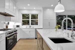Modern kitchen with white cabinets, stainless steel appliances, large island with sink, pendant light, and windows showing trees outside. Bright, clean, and spacious design with wood flooring.