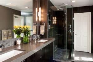 Modern bathroom with dark wood cabinets, double sinks, a glass-enclosed shower, gray tile accents, and a vase of yellow roses on the counter. White towels and bright lighting complete the clean, elegant look.