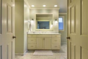 Spacious bathroom with cream cabinets, marble countertop, large mirror, wall sconces, towel, and a white rug on a marble floor, seen from an open doorway.