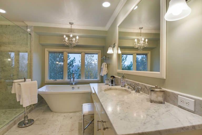 Elegant bathroom with a freestanding tub by large windows, marble countertop with dual sinks, wall-mounted lighting, a glass shower, chandeliers, and a view of trees outside.