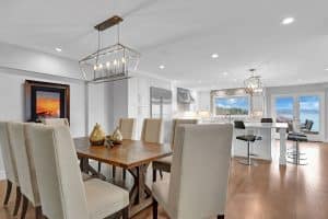 Modern dining and kitchen area with beige chairs, wooden table, pendant lights, white cabinets, and large windows showing ocean views and a sunset picture on the wall.