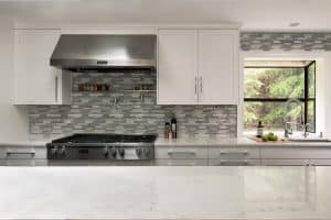 Modern kitchen with white cabinets, stainless steel range and vent hood, mosaic tile backsplash, and a large window above a sink with running water and green apples on the counter.