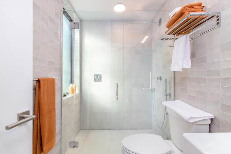 Modern bathroom with glass shower, white toilet, wall-mounted towel rack with orange and white towels, and a window. Orange towel hangs by the sink. Light gray tiles cover the walls and floor.