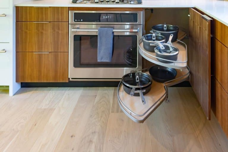 A kitchen corner cabinet with a pull-out shelf system holds pots and pans. The shelves swing out for easy access. An oven with a blue towel hanging on the handle is next to the cabinet.