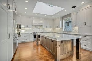 Modern kitchen with white cabinets, marble backsplash, stainless steel appliances, large center island with metallic accents, wood floor, and a skylight above providing natural light.