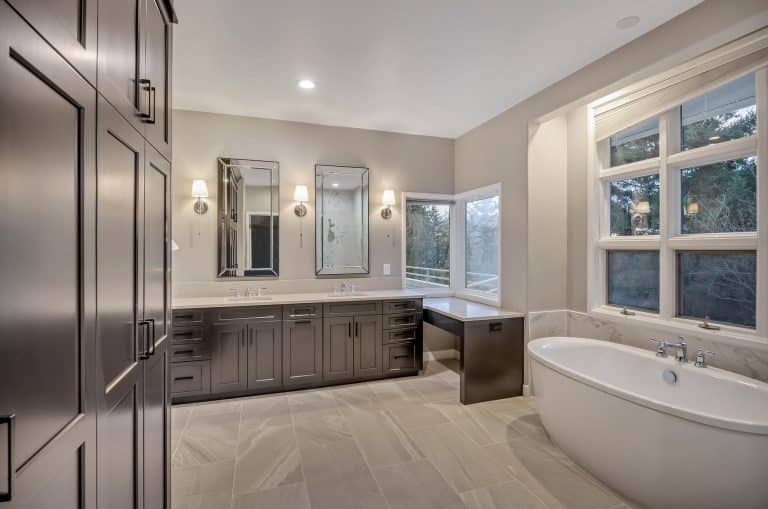 Modern bathroom with gray cabinets, double sinks, two mirrors, a soaking tub by large windows, and built-in vanity with a chair. Light walls and tiled floor, with wall sconces above the sinks and natural light through the windows.