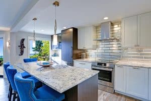 Modern kitchen with white cabinets, stainless steel appliances, marble countertop island, and blue barstools. Subway tile backsplash reflects light. Large windows reveal a scenic outdoor view.