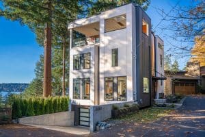 Modern three-story house with large windows, situated on a sloped lot near Lake Sammamish. Surrounded by tall trees and autumn leaves, the home features a garage at street level and balconies on upper floors.