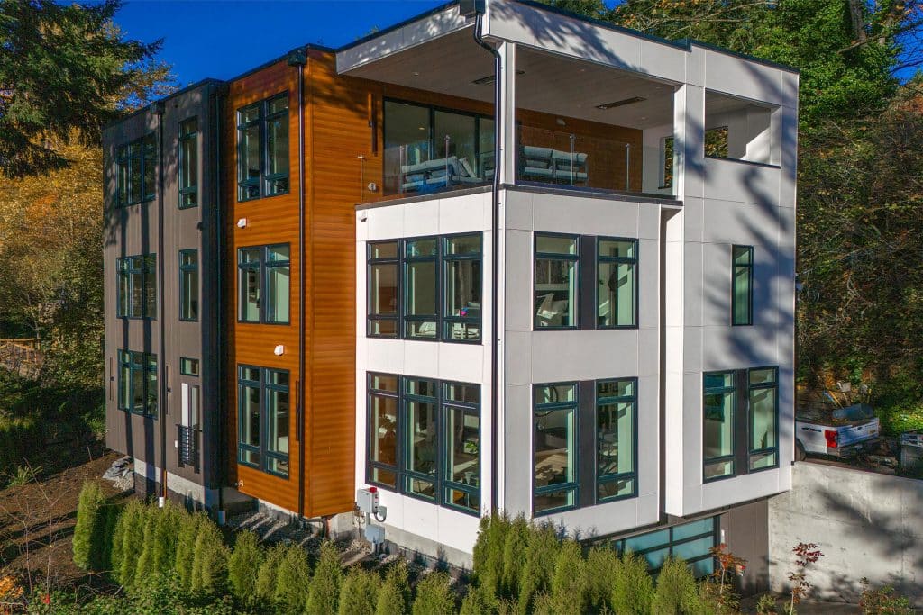 Modern three-story house with large windows, white and wood panel exterior, balconies, and a covered patio. Nestled near Lake Sammamish, the home is surrounded by evergreen trees and shrubs on a sunny day.