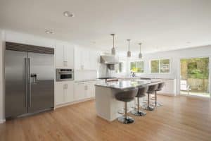 Modern kitchen with white cabinets, stainless steel appliances, a large island with four gray barstools, pendant lighting, and light wood floors. Large windows provide natural light and a view of the backyard.