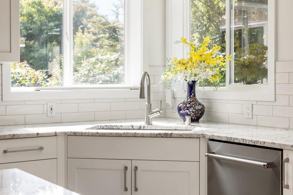 A bright kitchen with white cabinets, a marble countertop, a stainless steel sink and dishwasher, a vase of yellow and white flowers, and large windows letting in natural light.