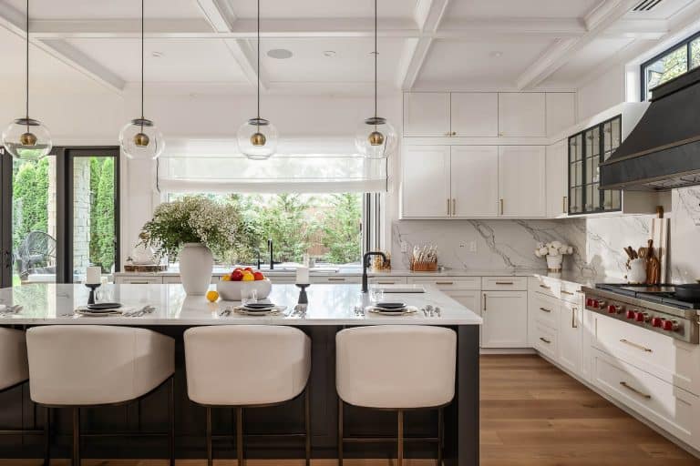 Modern kitchen with white cabinets, marble countertops, a large island with three white chairs, pendant lights, a gas stove with black hood, and large windows letting in natural light. Flowers and fruit decorate the island.