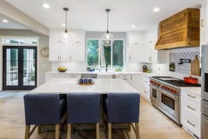 Modern kitchen with white cabinets, marble island, three blue chairs, wood range hood, stainless steel appliances, pendant lights, and a large window overlooking trees. Fruit bowls and decor items sit on the counters.