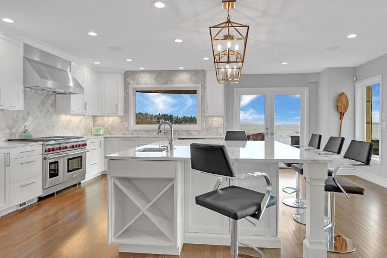 Modern white kitchen with island, black barstools, stainless steel appliances, pendant lighting, large windows, and a view of the ocean and beach. Perfect for anyone considering renovating your home, this space features a marble backsplash and wood floor that add elegant touches while helping you avoid costly mistakes often made during a home renovation.