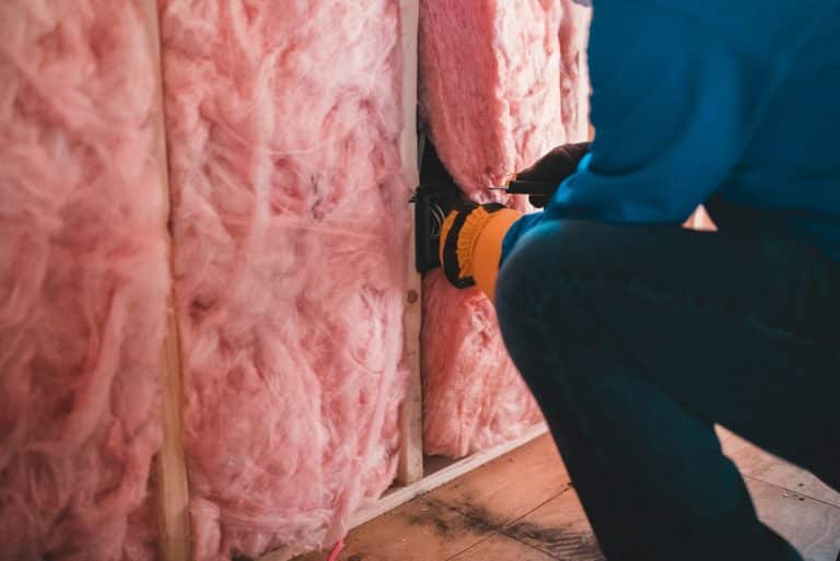 A person in gloves and a blue shirt installs or repairs an electrical outlet in a wall with exposed pink insulation, helping to ensure proper home insulation and potentially reduce energy bills.