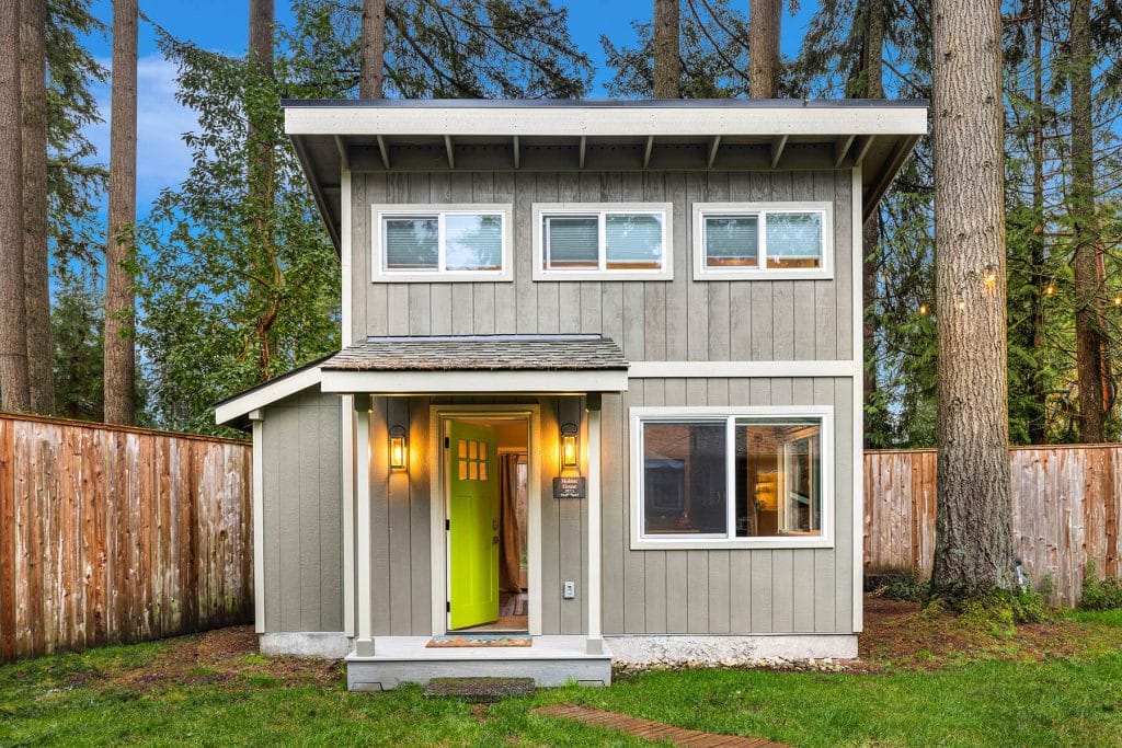 A small, modern gray house with a bright green door, lit porch lights, and multiple windows, surrounded by tall trees and a wooden fence, with grass and a stone path in front.