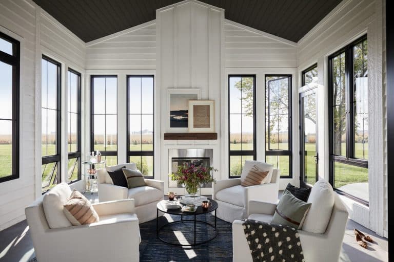Bright sunroom with large windows, four white armchairs around a round coffee table, a fireplace with art above it, and outdoor views of green fields. Homeowners will appreciate how the expansive windows not only fill the space with natural light but also help meet modern energy codes. Flowers and books decorate the space.