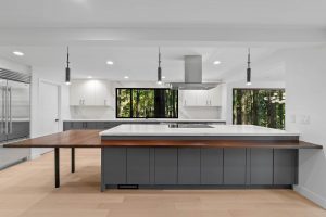 Modern kitchen with gray cabinets, a large island featuring white and wood countertops, pendant lights, stainless steel appliances, and large windows showing a green forest outside—perfect for any Bellevue home renovation or whole house remodel project seeking both style and functionality.