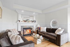 Bright living room with two brown sofas, a glass coffee table, and a lit fireplace. Family photos and candles decorate the white mantel. A round window and light wood floor add warmth and charm to this inviting Bellevue home, recently transformed as part of a Full House Remodel.