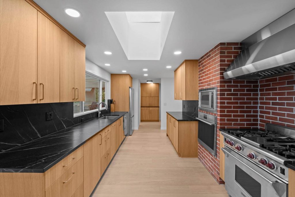 Modern kitchen with light wood cabinets, black countertops, stainless steel appliances, a gas stove, built-in microwave, and a red brick wall accent. Skylight and recessed lighting brighten the space.