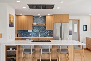 Modern West Seattle kitchen with light wood cabinets, blue tiled backsplash, stainless steel appliances, and a marble island with four gray cushioned barstools. Framed artwork decorates the walls.