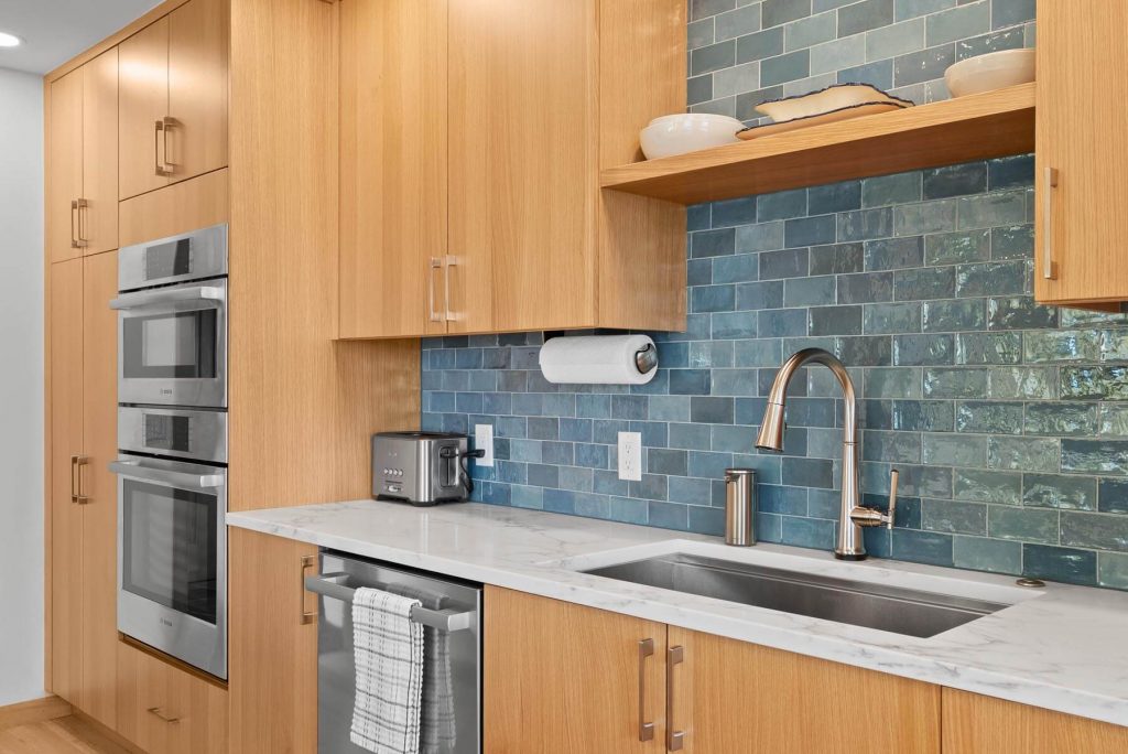 Modern West Seattle kitchen with wooden cabinets, marble countertop, stainless steel sink and faucet, toaster, double oven, and blue tile backsplash. A paper towel holder and dishes are also visible.