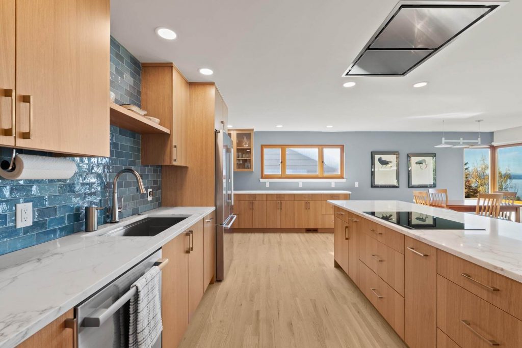 Modern West Seattle kitchen with wood cabinets, large island, blue tile backsplash, stainless steel appliances, and marble countertops. Large windows provide natural light, and a dining area is visible in the background.