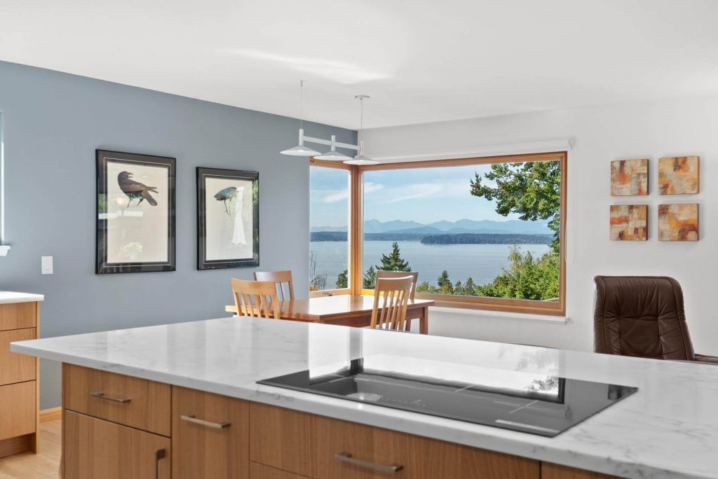 Modern West Seattle kitchen with marble island and built-in cooktop, wooden cabinets, dining area with two chairs, large window showing a lake and mountains, and framed bird art on a blue accent wall.