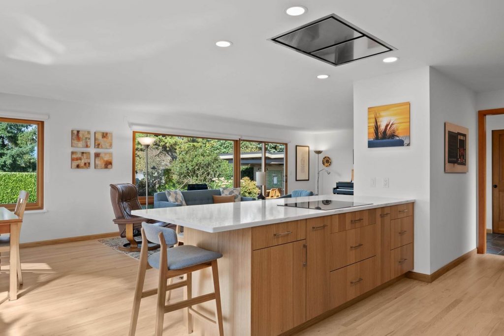Modern West Seattle kitchen with wooden cabinets, a white island countertop, bar stools, and large windows showing a green outdoor space. Living area with chairs and wall art is visible in the background.