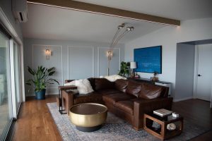 Modern living room with a brown leather sectional, gold round coffee table, plants, and wall art. Large window on the left lets in natural light. Light wood floors and contemporary lighting fixtures complete the space.